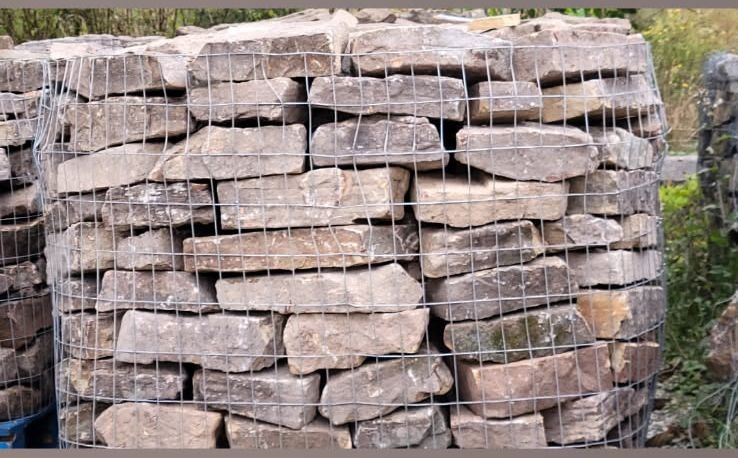 A round wire gabion cage filled with neatly stacked flat stone, set outdoors against green planting